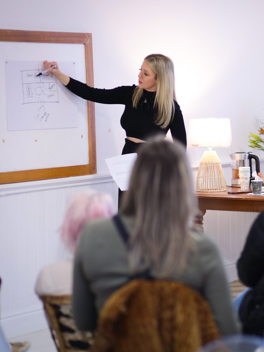 Hayley Michelle teaching a gathering of women, pointing to a floor plan drawn on the wall